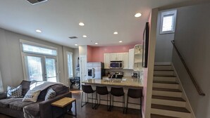 Una cocina moderna con armarios blancos, un refrigerador de acero inoxidable y un microondas. Hay una zona de bar con taburetes y una mesa de madera. La sala de estar tiene un sofá gris con cojines estampados y una mesa de centro. Se ve una escalera en el lado derecho.