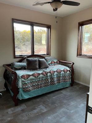 A bedroom with a bed, two windows, a ceiling fan, and a view of trees outside.