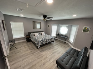 Ein Schlafzimmer mit einem Bett, einem Schreibtisch, einem Sessel, einem Deckenventilator und zwei Fenstern mit Jalousien.