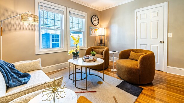 Un salon comprenant un canapé, un fauteuil, une table basse et une lampe. Il y a deux fenêtres avec des stores, une horloge et une porte.