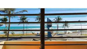 A view from a window with horizontal blinds, overlooking a beach with palm trees and the ocean.