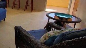 A room with a view of the ocean, a ceiling fan, a blue sofa, a wicker chair, and a small table with a green bowl.