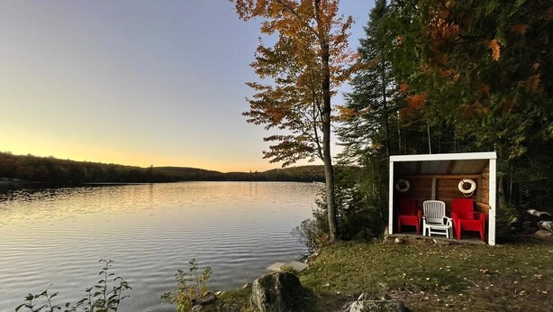 A lakeside cabin with a small porch, two chairs, and a view of the lake and forest. - Dawson Loop Inn - Private Lakefront Cottage (MONT)