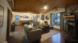 A spacious living room with a high ceiling, a fireplace, and a view through large windows. - Dawson Loop Inn - Private Lakefront Cottage (MONT)