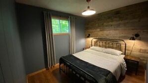 A bedroom with a bed, a nightstand, a lamp, a window with curtains, and a wooden wall. - Dawson Loop Inn - Private Lakefront Cottage (MONT)