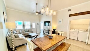 A spacious living room with a wooden dining table, white chairs, a sofa, and a ceiling fan.