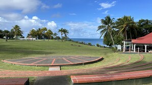 A grassy area with a circular stage, palm trees, and a building with a red roof. - Studio Ti Paradise Magnifique vue mer et Diamant (Sainte-Luce)