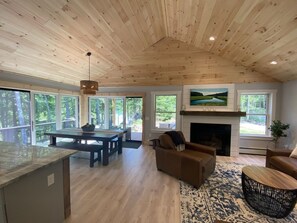 A spacious living area with a wooden ceiling, a fireplace, and large windows offering views of the outdoors. - Cozy 4 bedroom Lakehouse on Norway Lake (Norway)