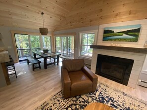 A spacious living room with a fireplace, a dining area with a table and benches, and large windows offering a view of the outdoors.