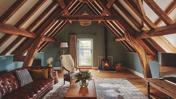 Ein gemütliches Dachzimmer mit freiliegenden Holzbalken, einem Ledersofa, einem Holztisch und einem rustikalen Kamin.