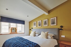 A bedroom with a large bed, a window with blinds, a bedside table, and wall-mounted artwork. - Hay Bale Cottage - Luxury Cottages (Worcester)