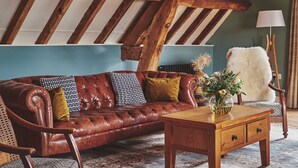 Ein gemütliches Loft mit freiliegenden Holzbalken, einem Ledersofa, einem Holztisch und einem flauschigen weißen Sessel.