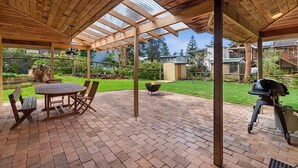 An outdoor patio with a wooden table and chairs, a grill, and a herringbone brick floor.