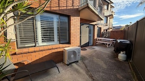 Een achtertuin van een woonhuis, met een terras, een tafel en stoelen, een grote airconditioning en een warmwaterboiler.