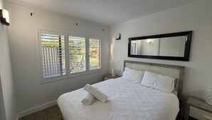 Een slaapkamer met een bed, nachtkastje, spiegel en raam met jaloezieën.