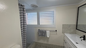 Badeværelse med bruser, toilet, håndvask og et vindue med skodder.