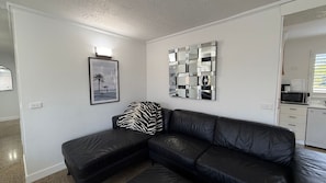 Un salon moderne avec un canapé en cuir noir, un coussin à motifs zébrés, une photographie encadrée représentant une plage et une œuvre d’art réfléchissante accrochée au mur.