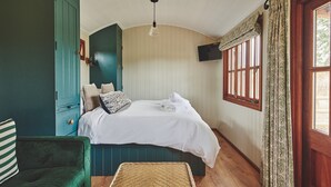 A bedroom with a bed, a green chair, a woven basket, and a window with curtains. - The Hangout Hut - Luxury Cottages (Worcester)
