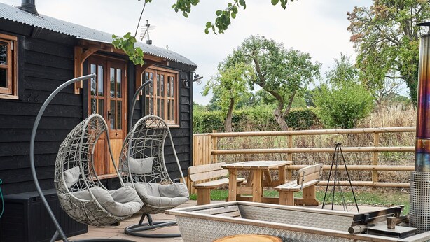 A wooden cabin with a swing chair, picnic table, and metal grill. - The Hangout Hut - Luxury Cottages (Worcester)