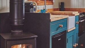 A kitchen with a wood-burning stove, wooden cabinets, a window with curtains, and a shelf with cups. - The Hangout Hut - Luxury Cottages (Worcester)