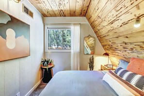 Une chambre avec un plafond en bois, un grand lit avec de nombreux oreillers, une table de chevet avec une lampe et une fenêtre donnant sur des arbres.