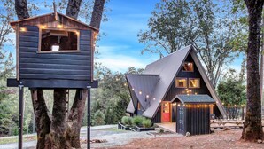A modern cabin with a large window and a treehouse attached to a tree. - Scenic A-frame - Hot Tub, Basketball Court, Treehouse, Outdoor theater! (Grass Valley)