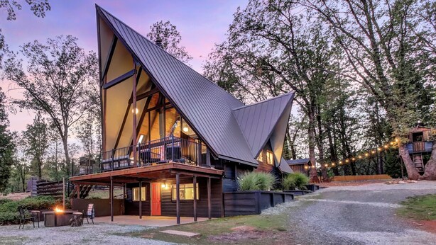 A modern, triangular-roofed cabin with a balcony, surrounded by trees. - Scenic A-frame - Hot Tub, Basketball Court, Treehouse, Outdoor theater! (Grass Valley)