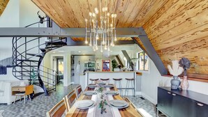A modern dining area with a long wooden table set for six, a spiral staircase, and a kitchen in the background. - Scenic A-frame - Hot Tub, Basketball Court, Treehouse, Outdoor theater! (Grass Valley)