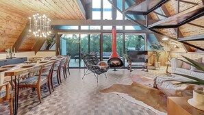 A spacious loft with a high ceiling, a dining area with a long wooden table, a cozy seating area with a fireplace, and large windows offering a view of the outdoors. - Scenic A-frame - Hot Tub, Basketball Court, Treehouse, Outdoor theater! (Grass Valley)