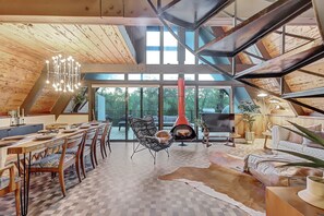 A spacious loft with a high ceiling, a dining area with a long wooden table, a cozy seating area with a fireplace, and large windows offering a view of the outdoors.