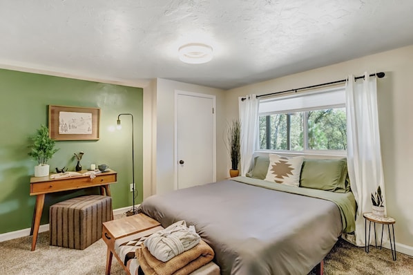 A bedroom with a bed, a desk, a chair, a window with curtains, and a door.