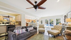 A spacious living area with a gray sofa, two armchairs, and a coffee table. A ceiling fan is visible, and the room has a large window offering a view of the outdoors.