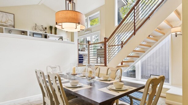 A dining area with a dark wooden table, white chairs, and a hanging light fixture. A staircase with wooden railings leads to an upper level.