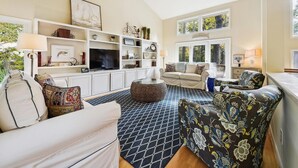 A living room with a sofa, armchairs, a coffee table, and a TV on a white wall. There are shelves with decorative items and a window with a view of trees.