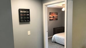 A hallway with a bed in the adjacent room, a wall-mounted light fixture, and a framed picture on the wall.
