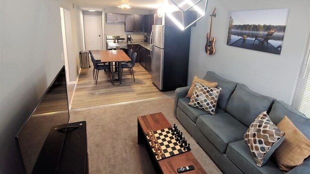 A modern living room with a grey sofa, a wooden coffee table, and a chessboard. There is a dining area with a table and chairs, and a kitchen in the background.