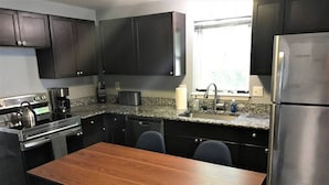 A modern kitchen with dark cabinetry, a stainless steel refrigerator, a granite countertop, and a wooden dining table with chairs.