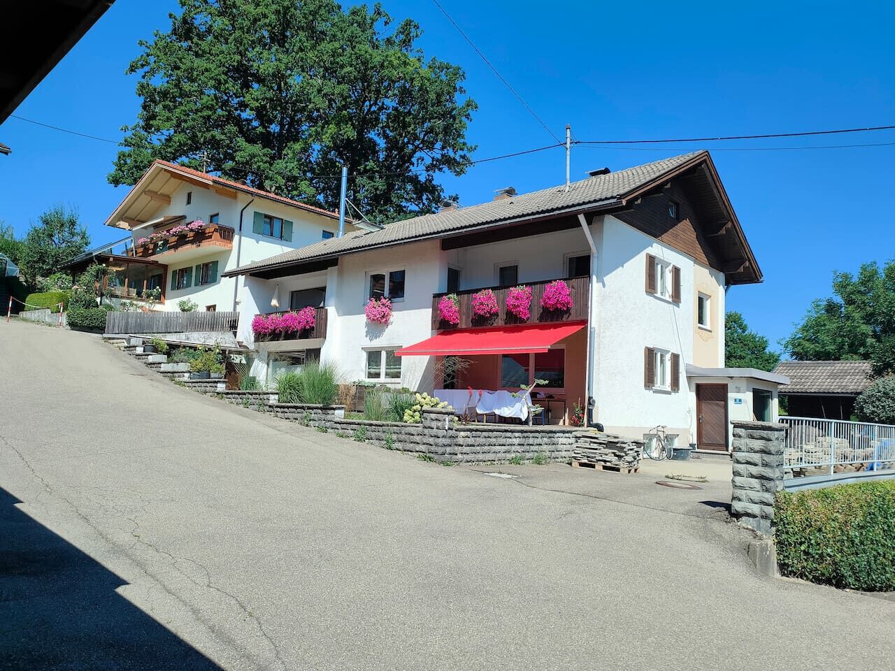 A two-story building with a red awning, a balcony with pink flowers, and a small attached structure.