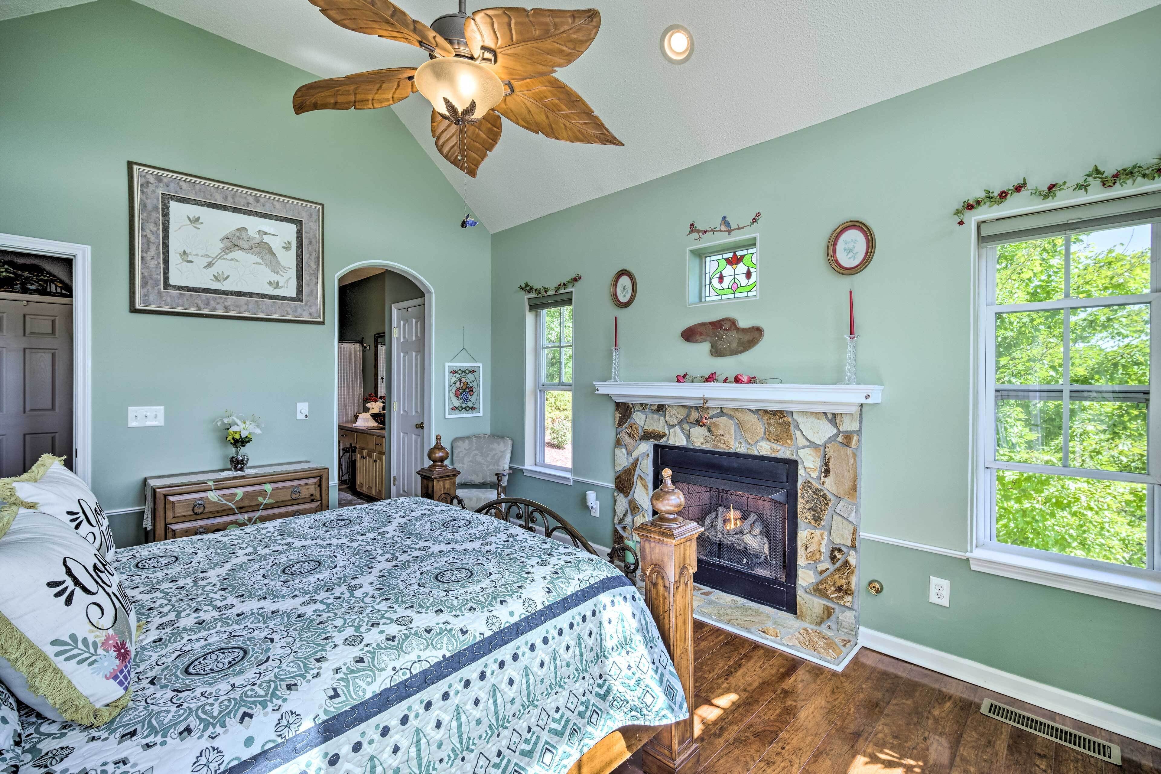 A bedroom with a large bed, a fireplace, a ceiling fan, and a window with a view of greenery.