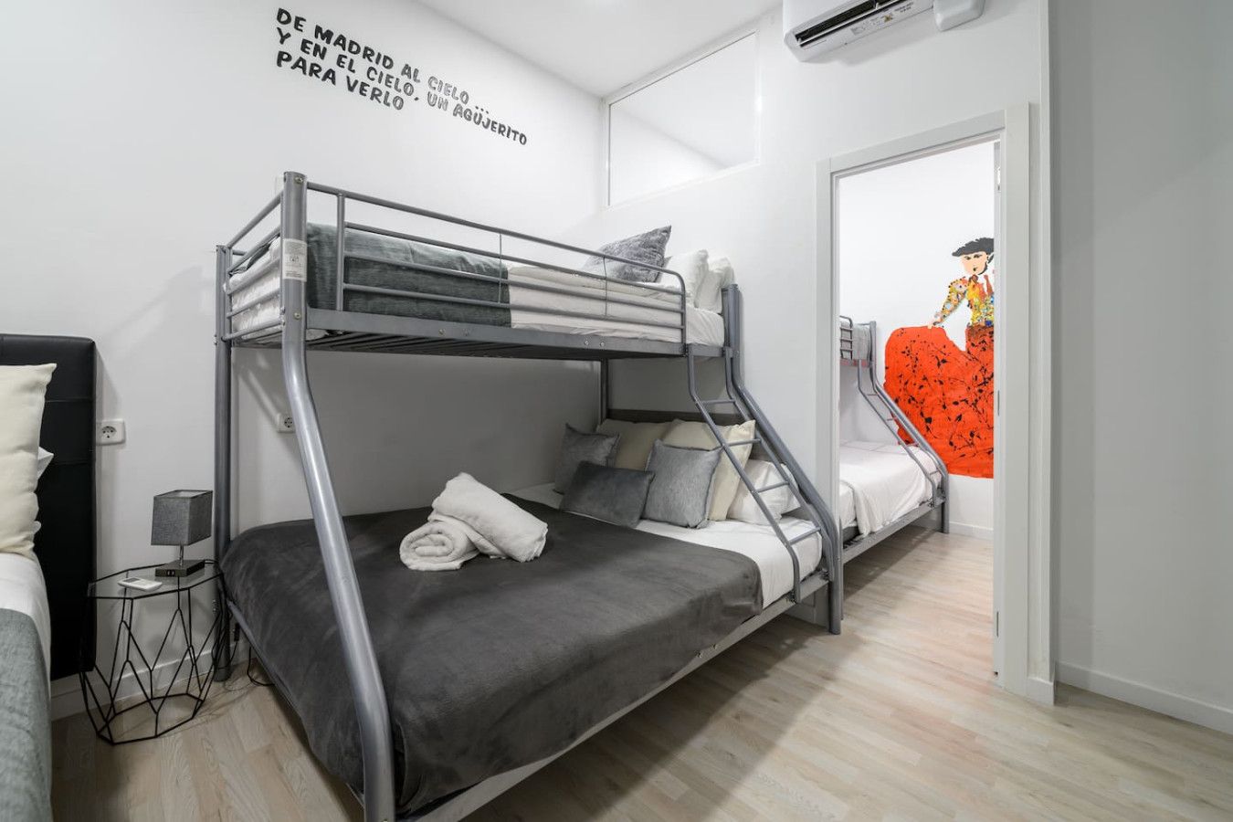 Habitación con literas, una cama a la izquierda y una litera a la derecha. Se observa una pared con una cita en español, una mesita con una lámpara y una puerta que da a otra habitación.