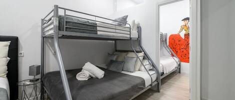 Habitación con literas, una cama a la izquierda y una litera a la derecha. Se observa una pared con una cita en español, una mesita con una lámpara y una puerta que da a otra habitación.