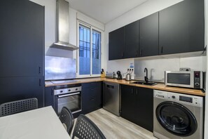 Une cuisine compacte avec des meubles de rangement sombres, un four à micro-ondes, un lave-linge et une fenêtre qui laisse entrer la lumière naturelle.