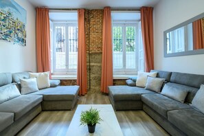 Una sala de estar con dos sofás grises, una mesita de centro y una planta en maceta. Hay ventanas con cortinas naranjas y una pared de ladrillo.