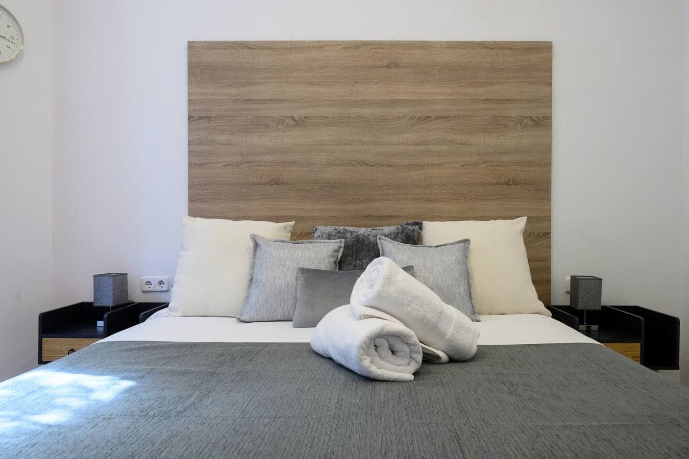 Ein ordentlich gemachtes Bett mit grauer Decke und aufgerollten Handtüchern, ein Holz-Kopfteil und zwei Nachttische mit Lampen.