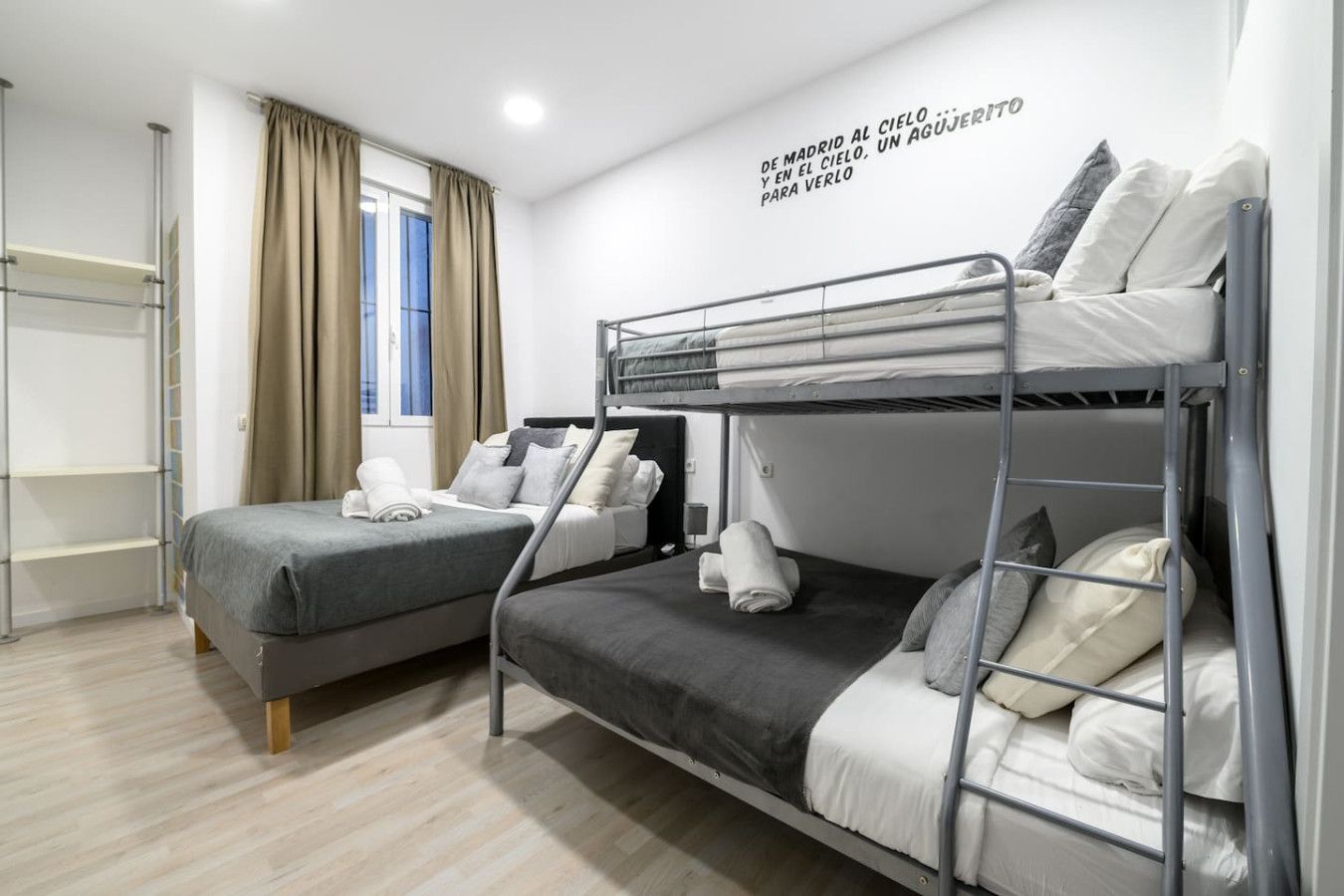Ein modernes Hotelzimmer mit Etagenbetten, einem Fenster mit Vorhängen, einem an der Wand montierten Regal und einer Deckenleuchte.