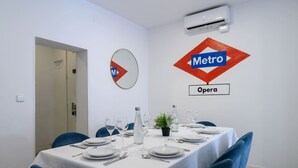 Un comedor con una mesa dispuesta para cuatro, mantel blanco, sillas azules y una planta en maceta.