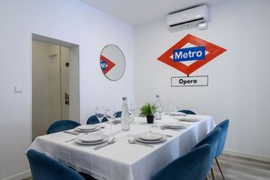 Een eethoek met een tafel gedekt voor vier personen, een witte tafelkleed, blauwe stoelen en een potplant.