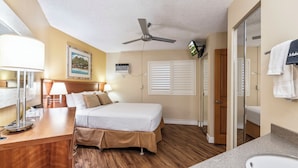 Habitación de hotel con cama, escritorio con lámpara, ventilador de techo, un cuadro en la pared y baño con lavabo y toallero.