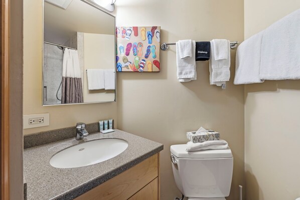 A bathroom with a sink, mirror, and toilet. There is a towel rack with multiple towels, a painting of flip-flops, and a makeup bag. - Maui Sunset Vacation Rentals A512 (Kihei)