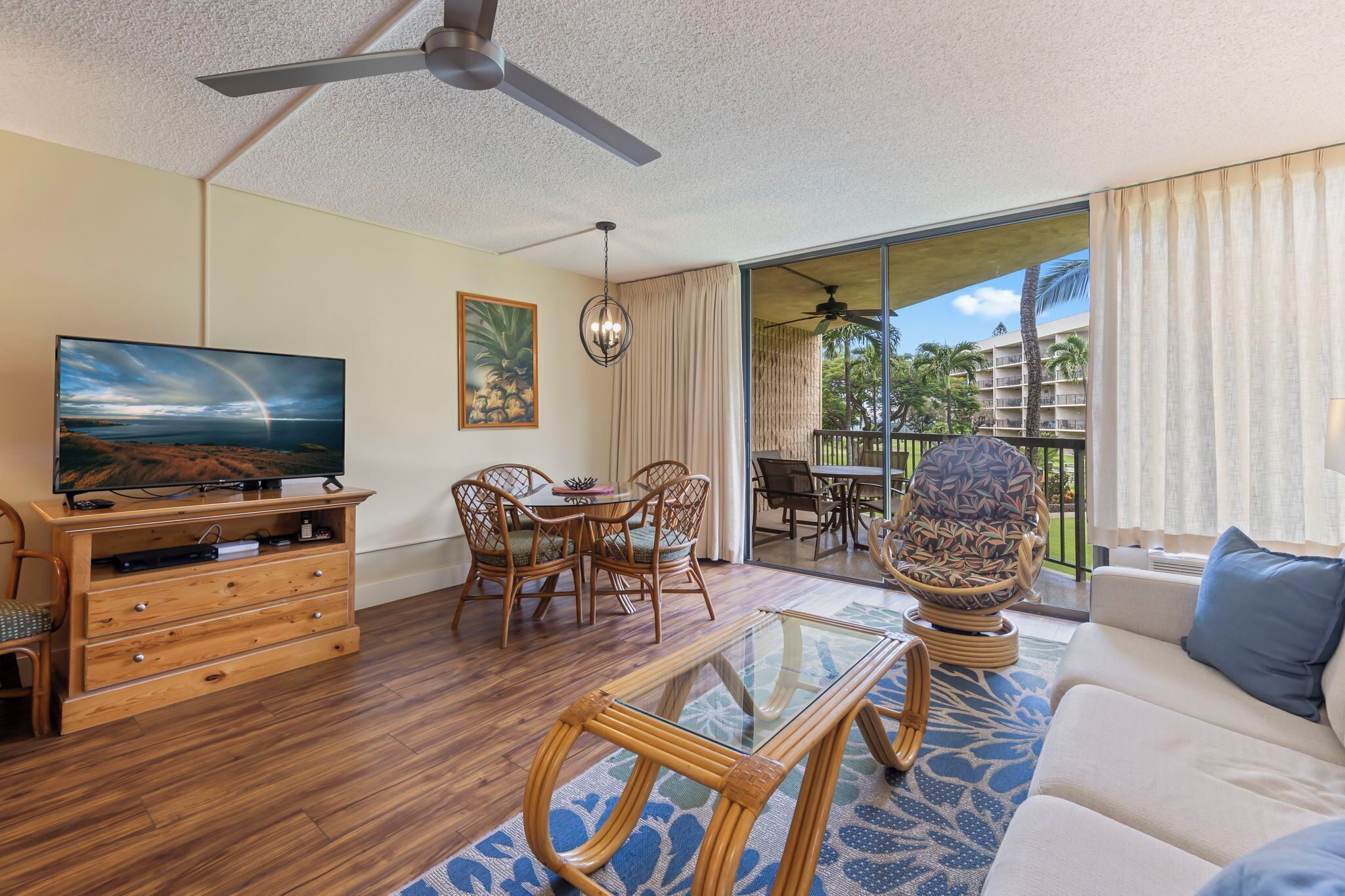 A living room with a television, wooden furniture, a glass coffee table, and a view of a balcony with outdoor seating.